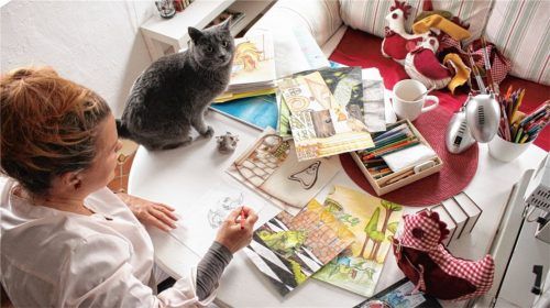 Kreativ mit Katze: Hier illustriert Autorin Cassandra May ihre Bücher, etwa das Fantasy-Märchen „Hallgard und das Vermächtnis von Origo“ und aktuell das Kinderbuch „Der Ostergockel Balduin“. Sie fertigt hier auch die lustigen Hühner zur Gockel-Geschichte als Stofftiere. Ihre Katze Emily schaut aufmerksam zu.Foto kla