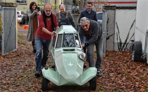 Klein und im Verhältnis gar nicht mal langsam: der Fend-Flitzer made in Rosenheim. Hier in Form einer Leihgabe aus Einbeck bei Hannover, hereingeschoben von (von links) Ausstellungsarchitekt Franz Putner, dem Oldtimer-Fachmann Wilfried Lebe aus Flintsbach, dem Rosenheimer Stadtarchivar und Ausstellungskurator Karl Mair und einem Fahrer des Transportunternehmens. Monika Hauser-Mair, die Leiterin der Städtischen Galerie Rosenheim, fotografiert.