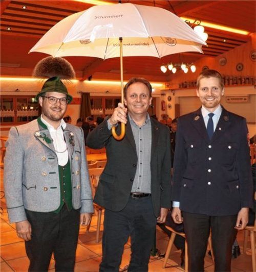 Als Zeichen seiner neuen Würde erhielt der Schirmherr der beiden Gründungsfeste 2023 in Frasdorf, Bürgermeister Daniel Mair (Mitte), bei der Jahresversammlung des Feuerwehrvereins in der Lamstoahalle einen großen weißen Schirm von den beiden Vorsitzenden Sebastian Graf (links) und Benno Voggenauer (rechts). Foto Rehberg