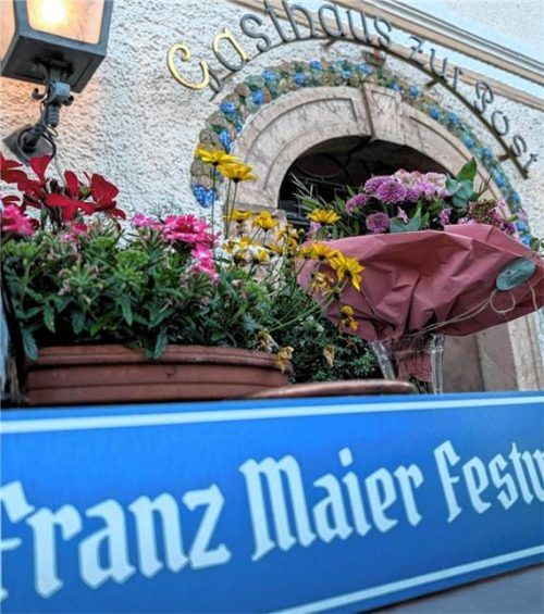 Das überreichte Schild mit der Aufschrift „Franz Maier Festwiese“ im Vordergrund, dahinter das Gasthaus zur Post mit Blumenarrangements. Hubert Hobmaier