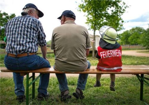 Ein kleiner Junge mit Feuerwehrhelm und Jacke sitzt neben zwei älteren Männern. Foto dpa