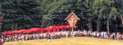 Zahlreiche Besucher sitzen an langen roten Biertischen und genießen das Waldfest. Im Hintergrund ist eine Bühne mit Auftritten zu sehen, umgeben von Bäumen und einer Berglandschaft. Foto Hoffmann