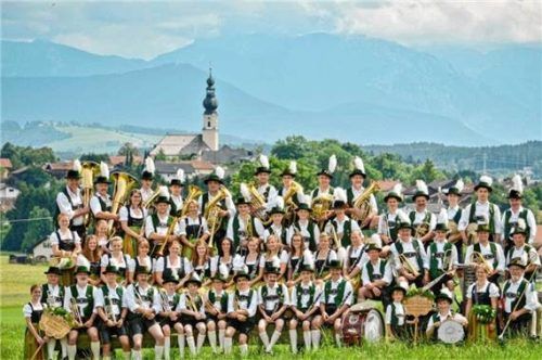 Eine große Gruppe von Musikern der Blaskapelle Traunwalchen posiert in traditioneller Tracht mit ihren Instrumenten vor einer malerischen Landschaft mit Bergen und einer Kirche. Foto Blaskapelle Traunwalchen