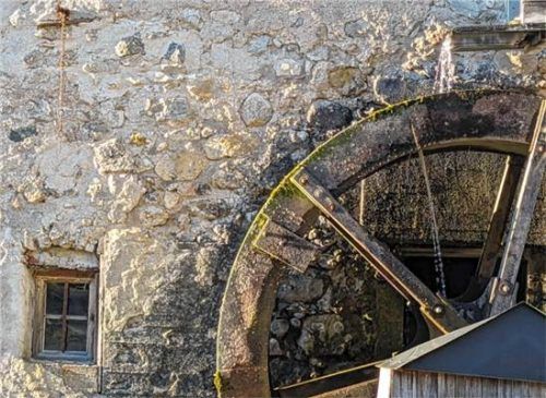 Ein Wasserrad an einer alten Steinmauer, das die Kraft des Mühlbachs nutzt.  Firmkäs